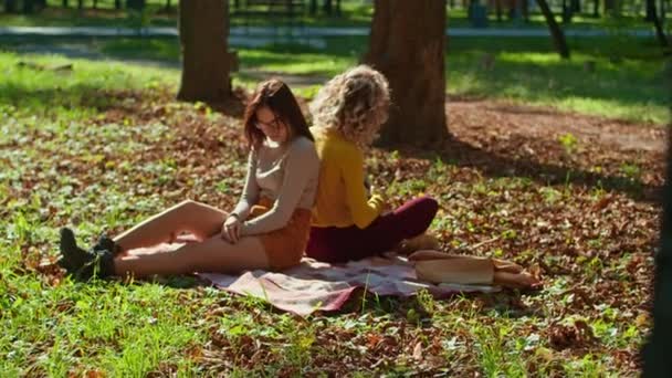 Két boldog fiatal nő jól érzi magát a városi parkban, az egyik lány gitározik, a másik a fák zenéjét és zaját élvezi. őszi nyári időszak. — Stock videók