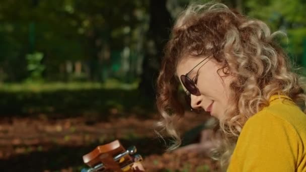 Δύο χαρούμενες νεαρές γυναίκες περνούν υπέροχα στο πάρκο της πόλης, το ένα κορίτσι παίζει κιθάρα, το δεύτερο απολαμβάνει τη μουσική και το θόρυβο των δέντρων. Φθινοπωρινή περίοδος. — Αρχείο Βίντεο