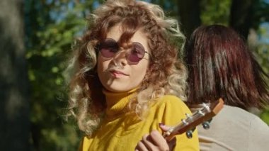 İki mutlu genç kadın şehir parkında harika vakit geçiriyor, bir kız gitar çalıyor, ikincisi ise müziğin ve ağaçların gürültüsünün tadını çıkarıyor. sonbahar yaz dönemi.