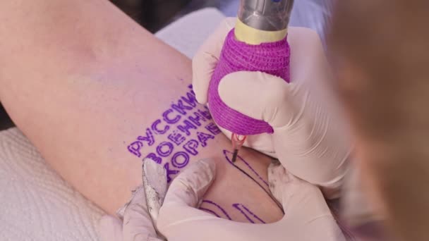 Närbild En professionell tatueringskonstnär gör en tatuering på en vuxen mans arm. tatueringen säger - gå ryska krigsfartyg. konflikt mellan Ukraina och Ryssland. krig 2022. — Stockvideo