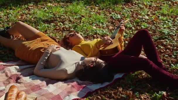 2人の幸せな若い女性が市内の公園で素晴らしい時間を過ごしている、 1人の女の子はギターを弾いている、 2番目の音楽と木のノイズを楽しんでいる。秋の夏. — ストック動画