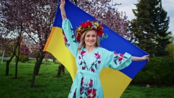 Щаслива жінка в вінку і з прапором України розважаються на тлі квітучих рожевих квітів сакури. Вихідні прогулюються з посмішкою на заході сонця. жінка і весна. свобода і незалежність . — стокове відео