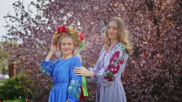 Dwie szczęśliwe kobiety w wieńcu na głowie radują się na tle kwitnących różowych kwiatów sakury. Weekendowy spacer z uśmiechem o zachodzie słońca. kobieta i wiosna. — Wideo stockowe