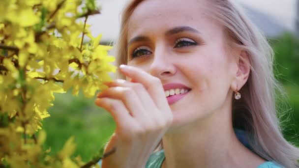 Oung blonde girl blows off a dandelion on a background of blooming pink sakura flowers. Weekend walk with a smile at sunset — Stock Video