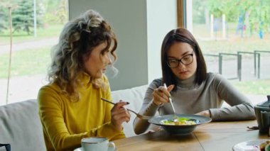 two young happy women enjoy delicious salad of chicken, tomato and croutons, eat it with a fork in a beautiful cool place in hot summer.