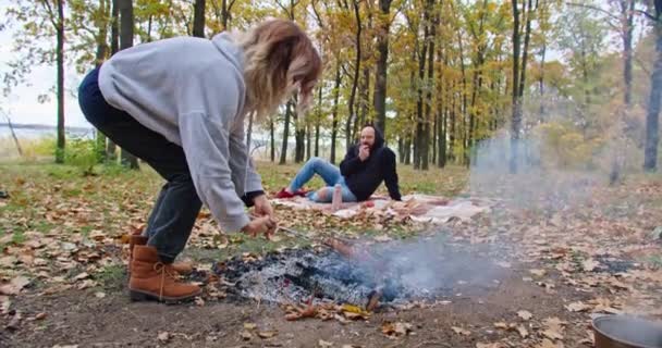 Happy girl fries sausages in nature, the guy is resting on a plaid, family autumn outdoor recreation. — Vídeos de Stock