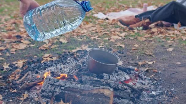 Happy young family couple resting in nature. the guy pours water into a cauldron for making tea. the girl is resting on a blanket in the autumn forest. — Vídeo de Stock