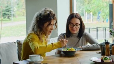 two young happy women enjoy delicious salad of chicken, tomato and croutons, eat it with a fork in a beautiful cool place in hot summer.
