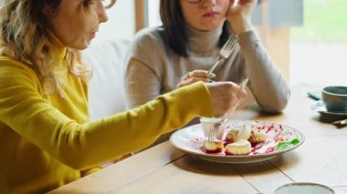 two young happy women enjoy delicious syrniki with jam, eat it with a fork in a beautiful cool place in hot summer.