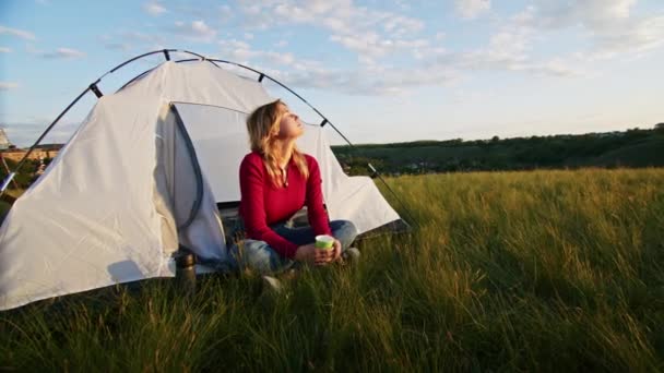 若い女性がハイキングの後の休息のために喜んで日没時に山の丘の上にテントを設置しました 熱いお茶を飲む女性 残りの1人は山の中で — ストック動画