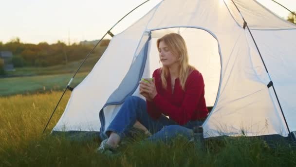若い女性がハイキングの後の休息のために喜んで日没時に山の丘の上にテントを設置しました 熱いお茶を飲む女性 残りの1人は山の中で — ストック動画