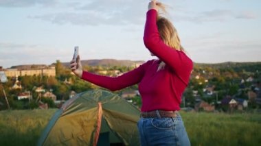 Genç bir kadın gün batımında dağ tepelerindeki bir telefonla yürüyüş sonrası rahatlamak için video iletişimi kurmaktan mutluluk duyar. Doğadaki diğer bir kişi, dağlarda.