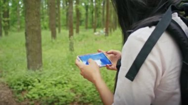 Sırt çantalı mutlu Asyalı bir kadın ormanda bir yol arıyor, telefon kullanıyor, görüntülü konuşuyor, haritaya bakıyor. yaz kampı