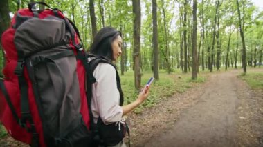 Sırt çantalı mutlu Asyalı bir kadın ormanda bir yol arıyor, telefon kullanıyor, görüntülü konuşuyor, haritaya bakıyor. yaz kampı