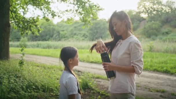 幸せなアジアの女性は娘と休暇を楽しんで森の道を歩き魔法瓶からお茶を飲む 家族のためのキャンプ 街の隣にある夏と都市公園 — ストック動画