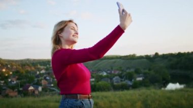 Genç bir kadın gün batımında dağ tepelerindeki bir telefonla yürüyüş sonrası rahatlamak için video iletişimi kurmaktan mutluluk duyar. Doğadaki diğer bir kişi, dağlarda.