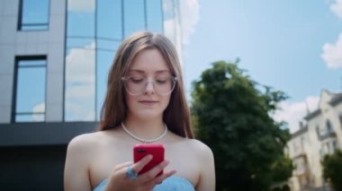 Mutlu bir kız telefonda video konuşması yapıyor ya da bir video izliyor, sokakta binanın arka planına karşı, rahat bir yaşam sürüyor ve yazın eğleniyor.
