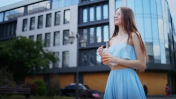 一个穿着蓝色衣服 戴着眼镜的快乐的女孩 靠着一栋大楼的背景 在街上喝着鸡尾酒 过着悠闲的生活 夏天过得很开心 — 图库视频影像