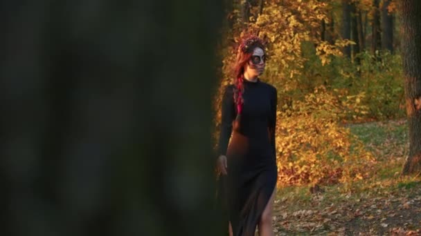 サンタ ムエルテの化粧をした若い女性は 日没時に森の紅葉を背景に黒い死のドレスを着て歩く 死者やハロウィーンのコンセプトの日 — ストック動画