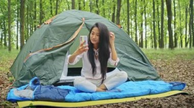 Mutlu Asyalı kadın tatilin tadını çıkarıyor, video görüşmesinde konuşuyor, telefonda film izliyor, Forest City Park 'ta çadırda dinleniyor. yaz kampı