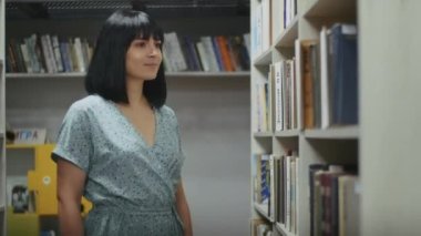 Mutlu ve hayalperest kadın kütüphanede kitap okuyor, raftan kitap seçiyor. Öğrenciler için kütüphanede öğrenim.