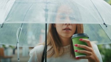 Neşeli kız yağmurlu bir havada şemsiyenin altında sokakta çay içer ve telefonda konuşur.
