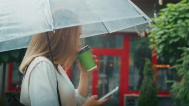 joyful girl drinks tea on the street under an umbrella in rainy weather, and speaks on the phone