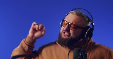 Close-up of a European podcast blogger smiling while broadcasting his live audio podcast in a studio using a microphone and headphones. Male radio host with glasses, podcast host or interviewer.