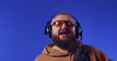 Close-up of a European podcast blogger smiling while broadcasting his live audio podcast in a studio using a microphone and headphones. Male radio host with glasses, podcast host or interviewer.
