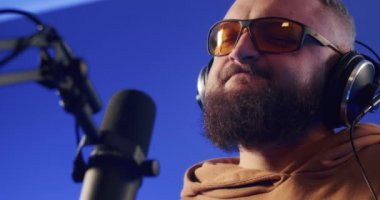 Close-up of a European podcast blogger smiling while broadcasting his live audio podcast in a studio using a microphone and headphones. Male radio host with glasses, podcast host or interviewer.