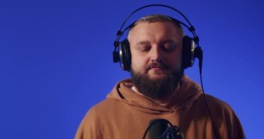 Close-up of a European podcast blogger smiling while broadcasting his live audio podcast in a studio using a microphone and headphones. Male radio host with glasses, podcast host or interviewer.