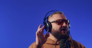 Close-up of a European podcast blogger smiling while broadcasting his live audio podcast in a studio using a microphone and headphones. Male radio host with glasses, podcast host or interviewer.
