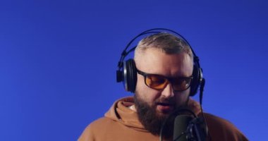 Close-up of a European podcast blogger smiling while broadcasting his live audio podcast in a studio using a microphone and headphones. Male radio host with glasses, podcast host or interviewer.