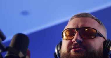 Close-up of a European podcast blogger smiling while broadcasting his live audio podcast in a studio using a microphone and headphones. Male radio host with glasses, podcast host or interviewer.