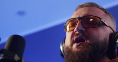 Close-up of a European podcast blogger smiling while broadcasting his live audio podcast in a studio using a microphone and headphones. Male radio host with glasses, podcast host or interviewer.