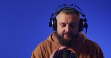 Close-up of a European podcast blogger smiling while broadcasting his live audio podcast in a studio using a microphone and headphones. Male radio host with glasses, podcast host or interviewer.