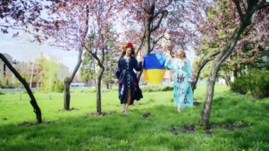 Ukrayna bayrağıyla çiçek açan pembe sakura çiçeklerinin arka planına sevinen iki mutlu kadın. Hafta sonu yürüyüşü, gün batımında bir gülümsemeyle. Kadın ve bahar