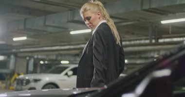 Siyah takım elbiseli, beyaz gömlekli ve kırmızı kravatlı güzel bir sarışın etrafına bakıyor, yeraltı otoparkında sırıtıyor, sinematik bir çekim, yavaş çekim.