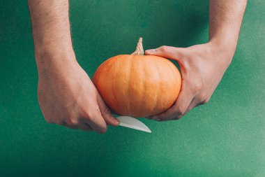 Erkeklerin elinde taze balkabağı ve bıçakla yaratıcı bir Cadılar Bayramı fikri. Asgari kesim ve zanaat konsepti, tatil sezonu estetik görselleştirme. Huysuz koyu yeşil arkaplan.