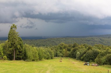 Yeşil dağ manzarası. Traktör çayırdan saman topluyor. Karpat Dağları, Ukrayna, Doğu Avrupa. Ormanın üzerinde ufukta gök gürültülü fırtına var..