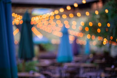 Odaklanmamış açık hava verandası. İnsanlar şenlikli bir atmosferde dinleniyor. Işıklar lambalardan geliyor, sarı bokeh etkisi.