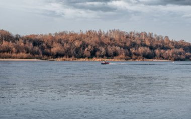Sonbaharda çıplak ağaçların ve fırtınalı bulutların arka planında küçük bir motorlu tekne olan bir nehir yüzeyi..