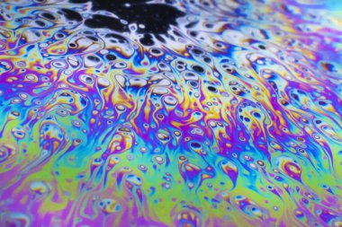 Bright green, pink, purple, blue and orange pastel colours swirl around intense black and white dots patterns on the surface of a soap bubble. 