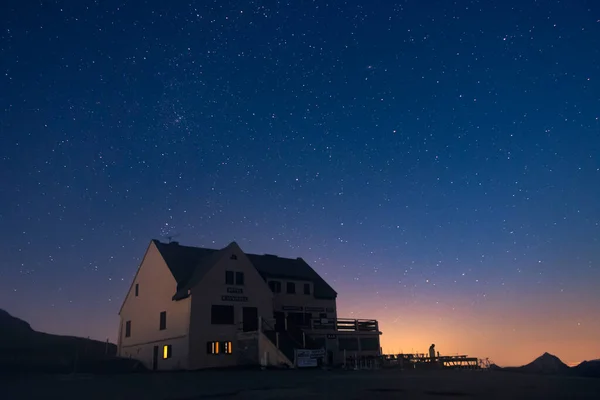 A pub stands alone shortly after the sun sets, with orange skyglow on the horizon as dark blue skies take over, twinkling with stars. Taken at Col dAubisque, a mountain pass in the Pyrenees