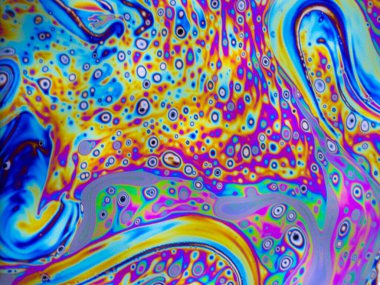Bright green, pink, purple, blue and orange pastel colours swirl around intense black and white dots patterns on the surface of a soap bubble. 