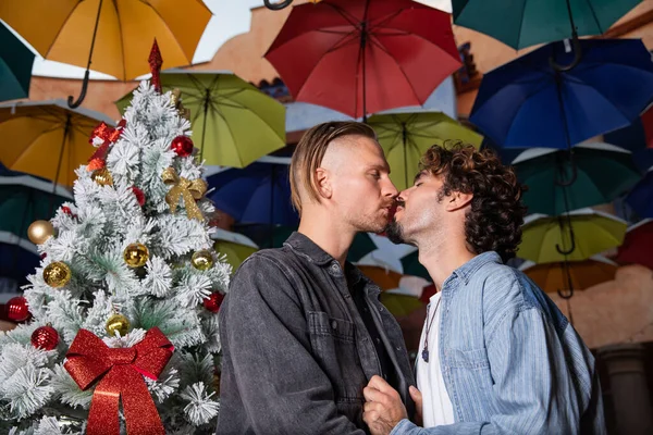 同性伴侣们分享着拥抱和亲吻的浪漫时刻 今天是圣诞节 他们站在一棵挂着五彩缤纷彩虹雨伞的圣诞树旁 同性恋夫妇Lgbtq概念 — 图库照片