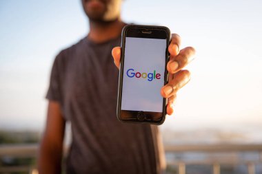 Adamın biri ekranında Google logosu olan bir akıllı telefon tutuyor. Google, internet hizmetleri ve ürünleri konusunda uzmanlaşmış bir Amerikan çokuluslu teknoloji şirketidir.