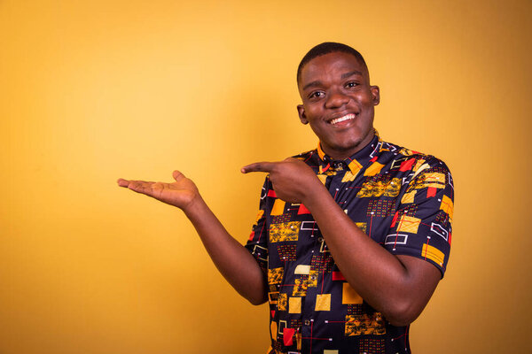 Young smiling African boy isolated on orange background, wearing African patterned shirt, his finger points to the space on his hand, advertising photo with copy space
