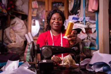 Afrikalı bir terzi elinde bir bobbin tutuyor ve terzi dükkanında çalışıyor, mutlu ve gülümsüyor.