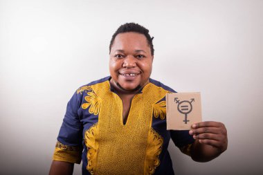 Trans sembollü bir karton taşıyan Afrikalı bir trans kişinin stüdyo portresi. Geleneksel bir Afrika elbisesi giyiyor.